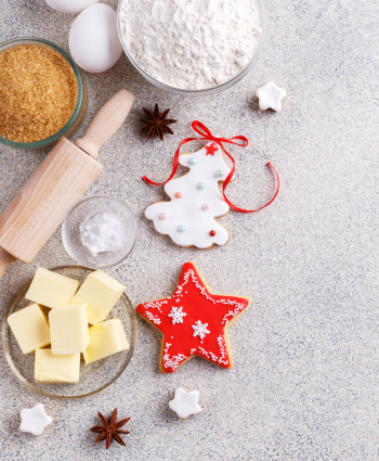Baking scene with flour, eggs, butter, brown sugar, and a rolling pin. Decorated Christmas cookies in tree and star shapes, surrounded by spices.