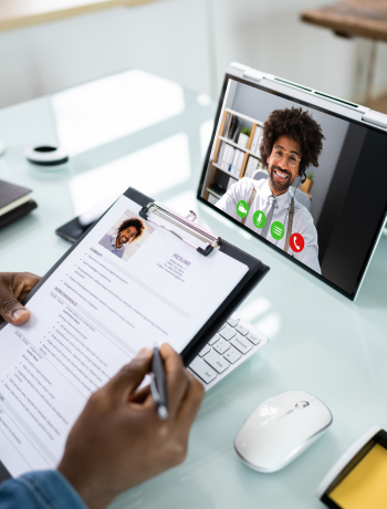 A person holding a resume is engaged in a video call on a laptop. The screen shows a smiling individual in a virtual meeting.