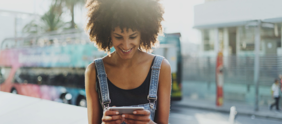 Smiling woman with curly hair wearing denim overalls looks at her phone. She's outdoors in a sunny urban setting with a colorful bus in the background.