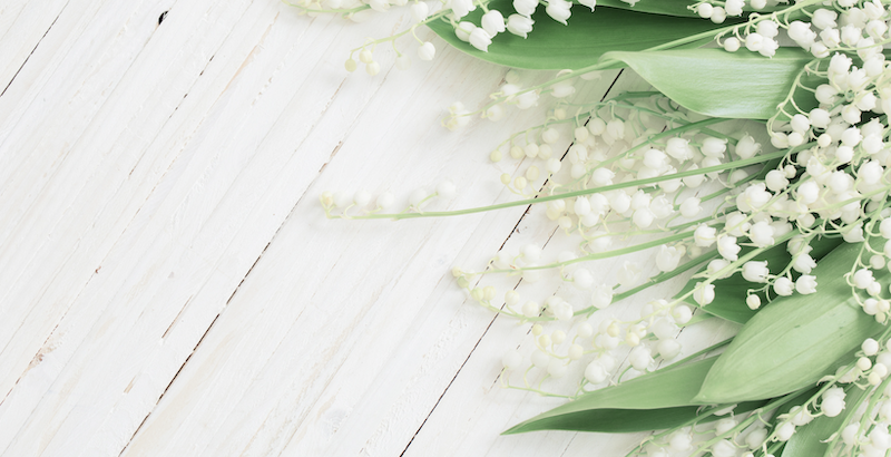Delicate lilies of the valley with green leaves lay on a light wooden surface, creating a serene, fresh, and elegant springtime feel.