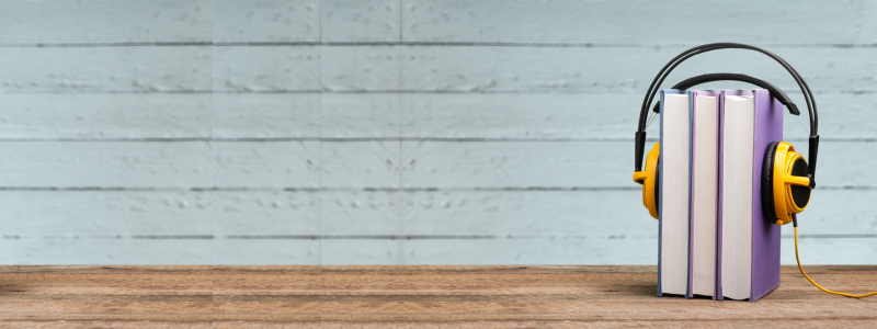Books stacked upright with a pair of yellow headphones resting around them on a wooden surface. Light blue wooden plank background conveys a cozy, calm tone.