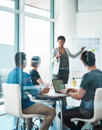 A professional woman presents at a meeting, pointing to a flip chart. Three colleagues sit at a table with laptops, focusing attentively. Bright, modern office.