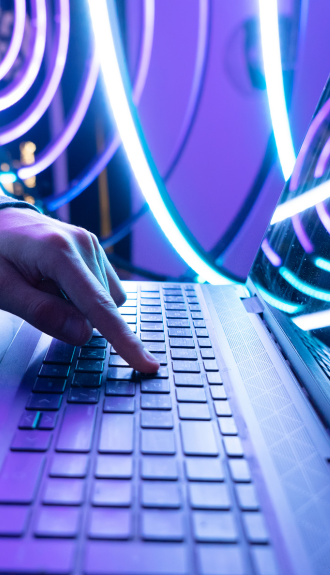A hand types on a laptop keyboard under vibrant neon lights, creating a futuristic ambiance with swirling blue and purple patterns.
