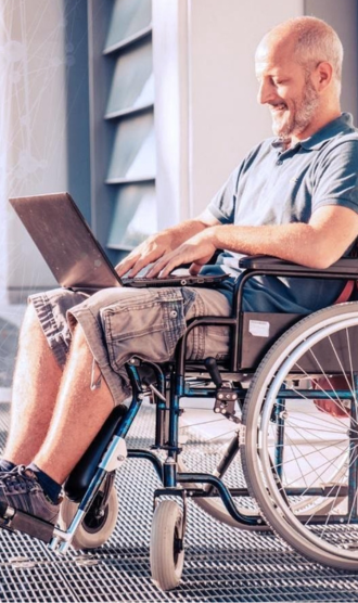 Man in a wheelchair smiles while typing on a laptop. He's wearing a blue polo and shorts, seated outside on a sunny day. The mood is positive and focused.