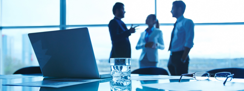 A conference room table with a laptop, documents, a glass of water, and glasses in focus. In the blurred background, three businesspeople are conversing.