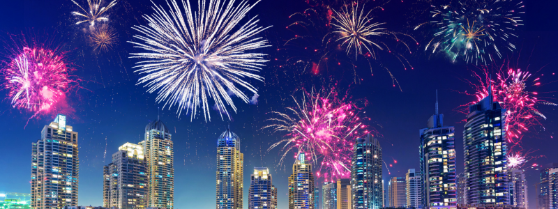 Fireworks explode over a modern city skyline at night, with bursts of red, pink, and white lights. The scene conveys celebration and excitement.