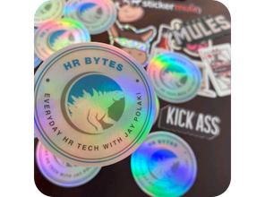 Holographic stickers on a table featuring 'HR Bytes,' a podcast-themed design with text 'Everyday HR Tech with Jay Polaki.' Background has more stickers.
