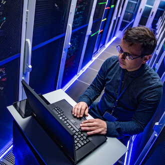 A person using a laptop on a small table in a dimly lit server room. They are surrounded by tall server racks with blue lighting. The individual is wearing glasses and a dark shirt.