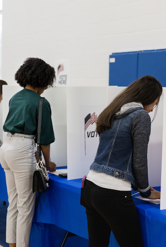 Two people, one wearing a green shirt and beige pants, the other in a denim jacket and black pants, are standing at voting booths with privacy screens displaying an American flag and the word VOTE.