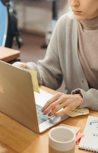 Person in a gray cardigan and turtleneck using a laptop at a wooden desk, with sticky notes attached. A notebook with a grid pattern and a cup are nearby. The scene suggests a work or study environment.
