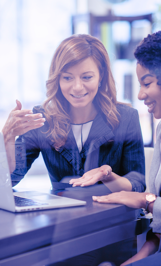 Two women seated at a table discussing something on a laptop. One woman has long hair and gestures with her hand, while the other, with short curly hair, is smiling and looking at the screen. Both wear blazers, suggesting a professional setting.