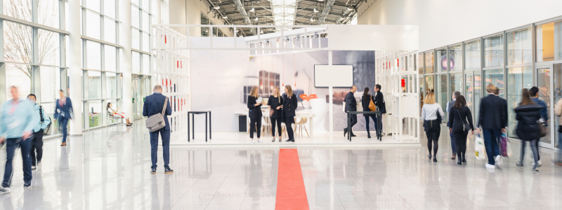 Blurred people walk past a modern, bright trade show booth with a red carpet. The atmosphere is busy and professional.