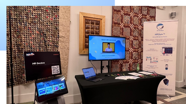 Exhibition booth with two screens showing digital content, a laptop, brochures, and a banner. The setting appears modern and professional.