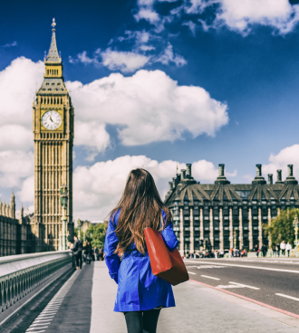 A woman in a blue coat walks towards Big Ben on a sunny day, with fluffy white clouds in a bright blue sky. She carries a red bag over her shoulder.