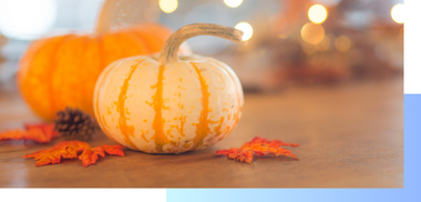 Small white and orange striped pumpkin on a wooden table with scattered autumn leaves and blurred bokeh lights, evoking a cozy fall ambiance.