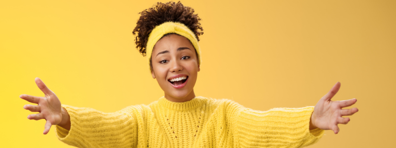 A joyful woman in a yellow sweater and headband extends her arms toward the camera, smiling broadly. The background is a matching warm yellow shade.